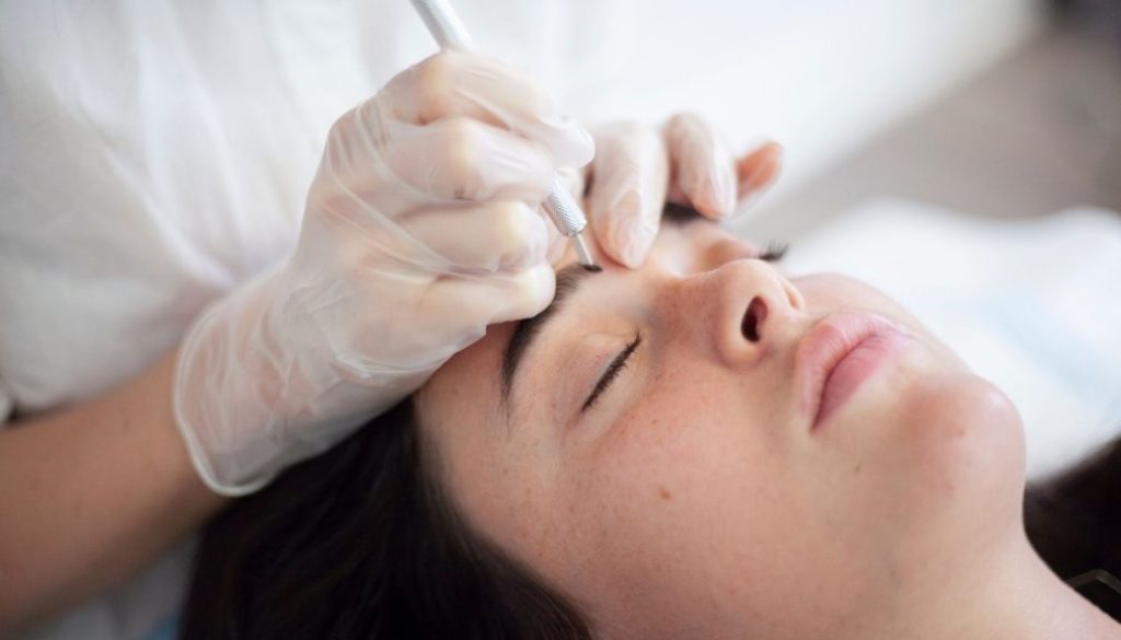 Close-up of a Blink Beauty Bar client receiving a microblading eyebrow treatment, with a gloved brow artist carefully applying hairstrokes.