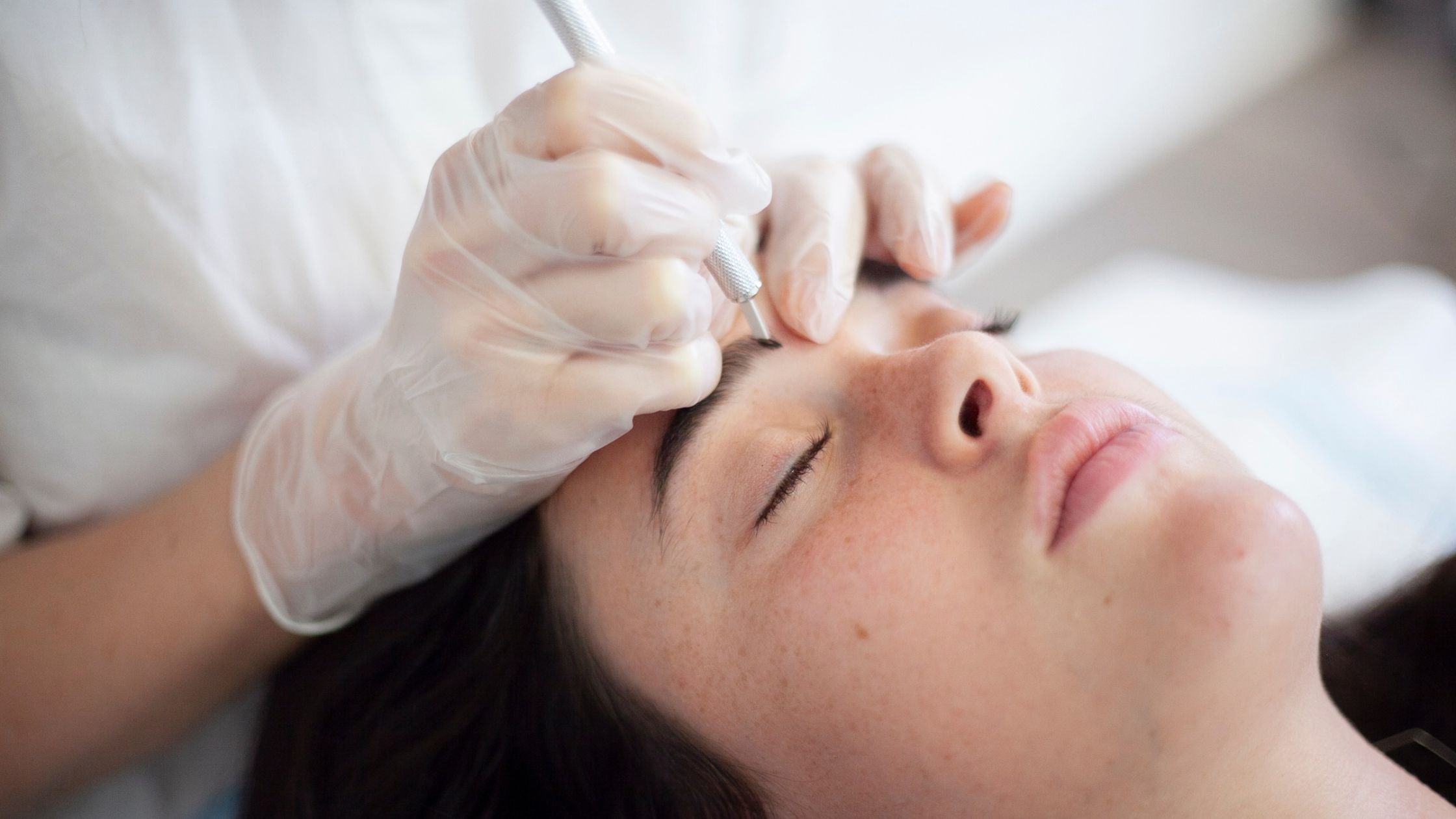 Close-up of a Blink Beauty Bar client receiving a microblading eyebrow treatment, with a gloved brow artist carefully applying hairstrokes.