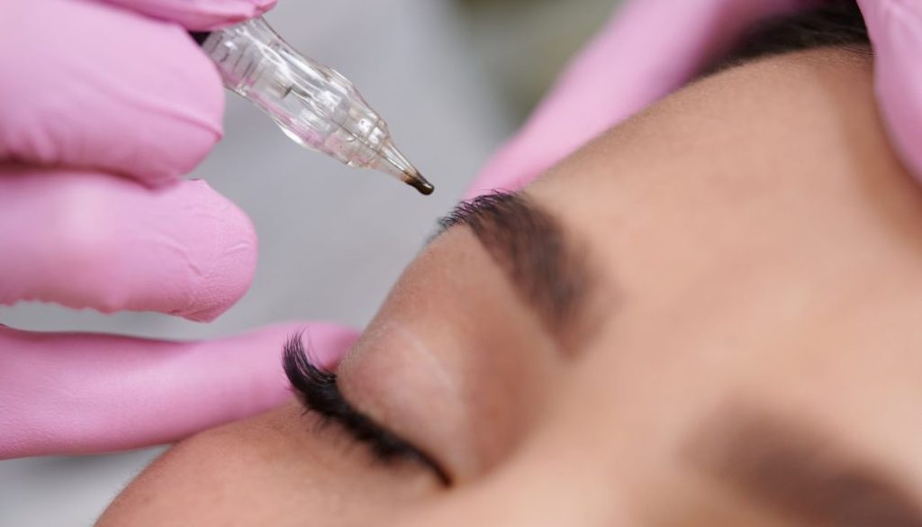 Close-up of a permanent makeup machine applying pigment to an eyebrow during a cosmetic brow tattooing treatment.