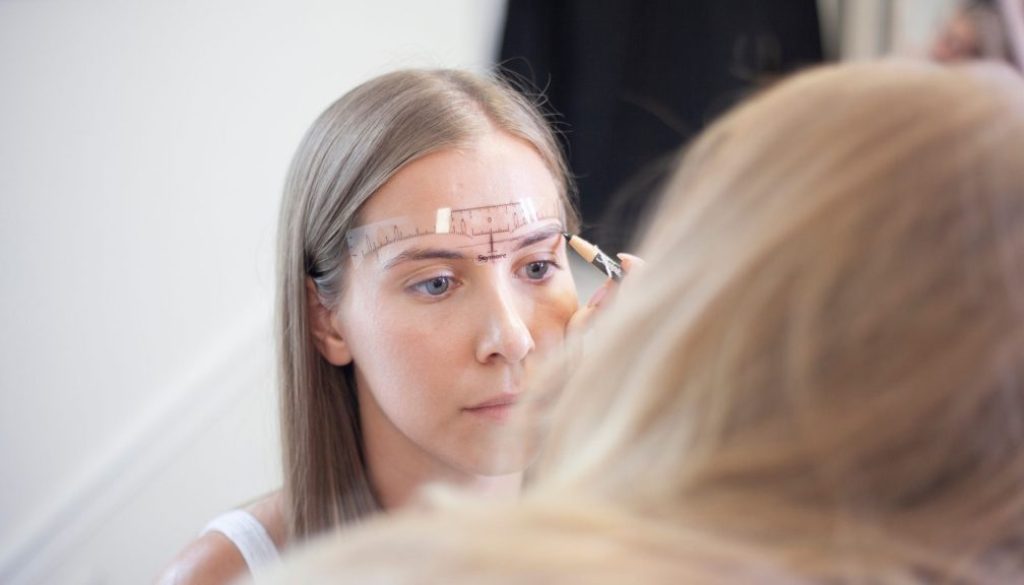 Blink Beauty Bar founder Ashley Schuh measuring and mapping a client's eyebrows with a brow ruler and pencil before a permanent makeup treatment.